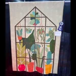 Greenhouse and plant themed quilt on a white background hanging at a quilt show with a ribbon