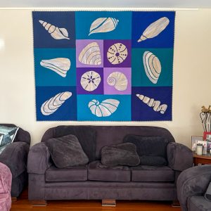 Blue, Purple, and off-white seashell quilt made with the Shoreline Shells Block of the Month pattern hanging over a couch