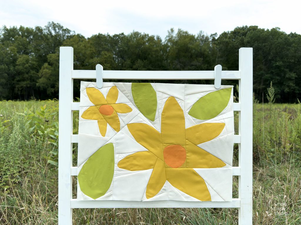 quilt block with yellow and orange flowers and green leaves on a ladder in a field