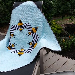 Bee-themed quilt featuring modern pieced bees and geometric shapes made with white, black, yellow fabrics on an aqua background draped over a chair outside