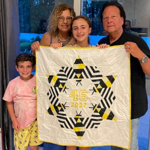 Four people holding up a modern bee quilt with graphic yellow, gray, and black bee blocks arranged on a light background.